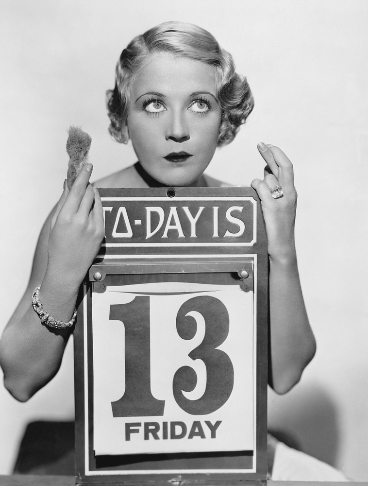 Black and white photo wit woman holding a calendar with Friday the 13th highlighted and she is crossing her fingers.