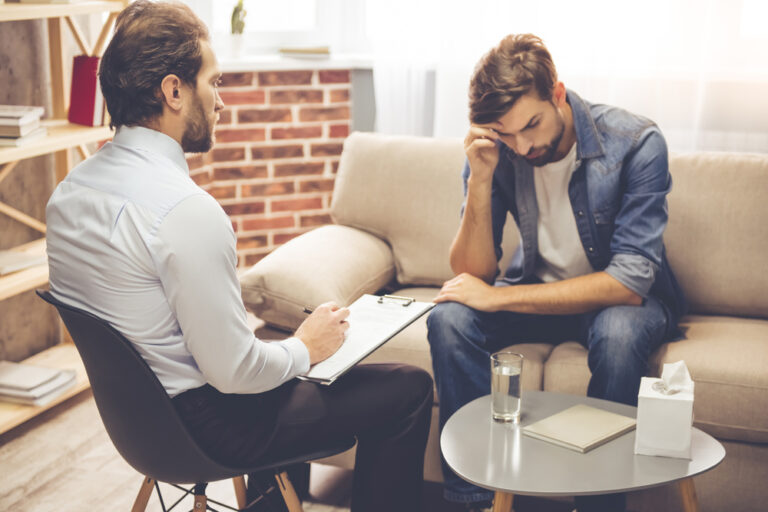 Male Counselor with pen and pad, facing a stressed young man on a couch.