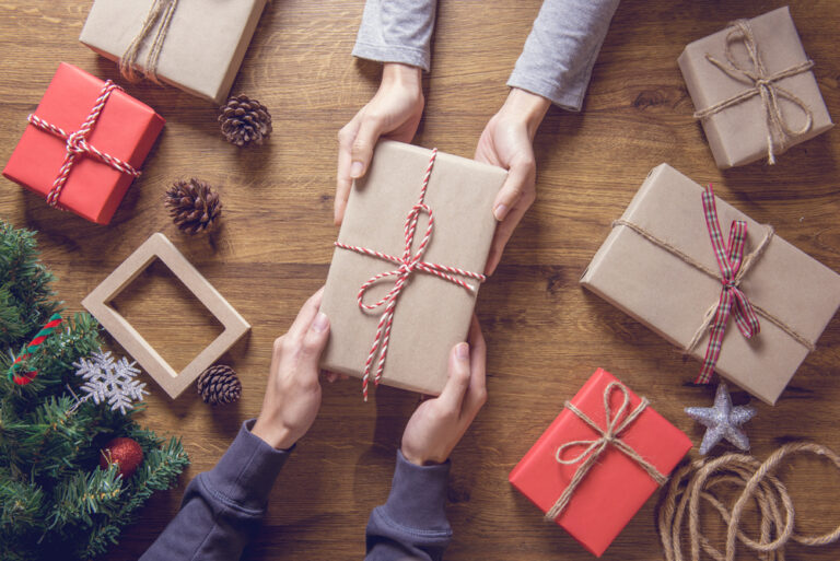 To hands giving wrapped gifts to each other, on a wood table with Christmas decorations.