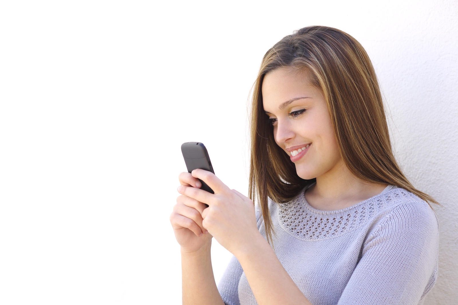 Happy woman texting on a smartphone looking at phone on a white wall