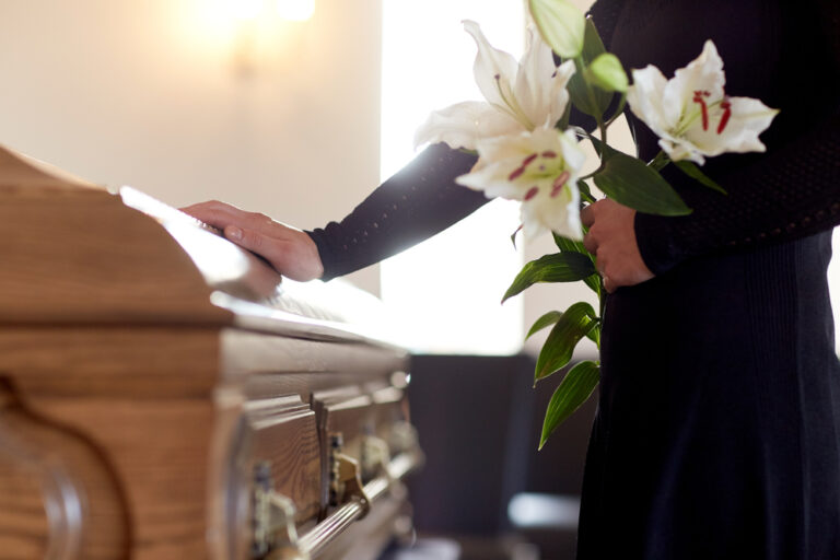Woman in black dress standing with hand on casket in funeral home, holding white lilies.