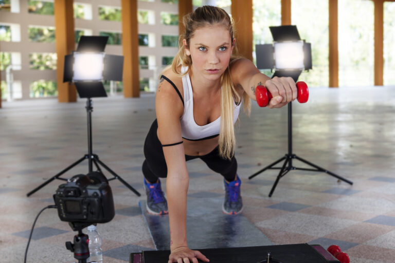 Young woman doing a plank with a dumbbell in one hand, while cameras and lights surround her for her social media.